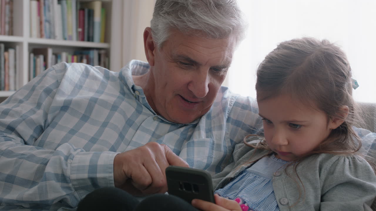 happy grandfather showing little girl how to use smartphone teaching curious granddaughter modern technology intelligent child learning mobile phone sitting with grandpa on sofa