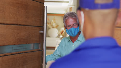 Delivery-man-delivering-package-to-senior-caucasian-woman-wearing-face-masks-at-home