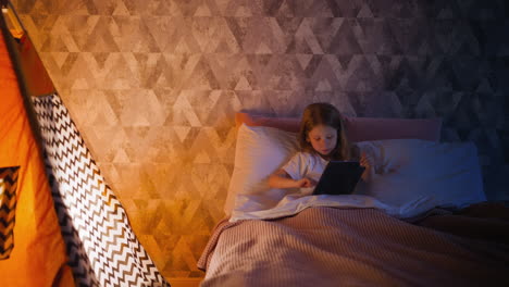 kid girl touches screen of tablet lying in bed by play tent