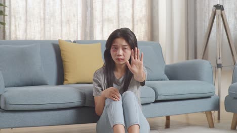 young asian woman with bruise on body looking into camera showing hand sign to stop violence while sitting on the floor at home