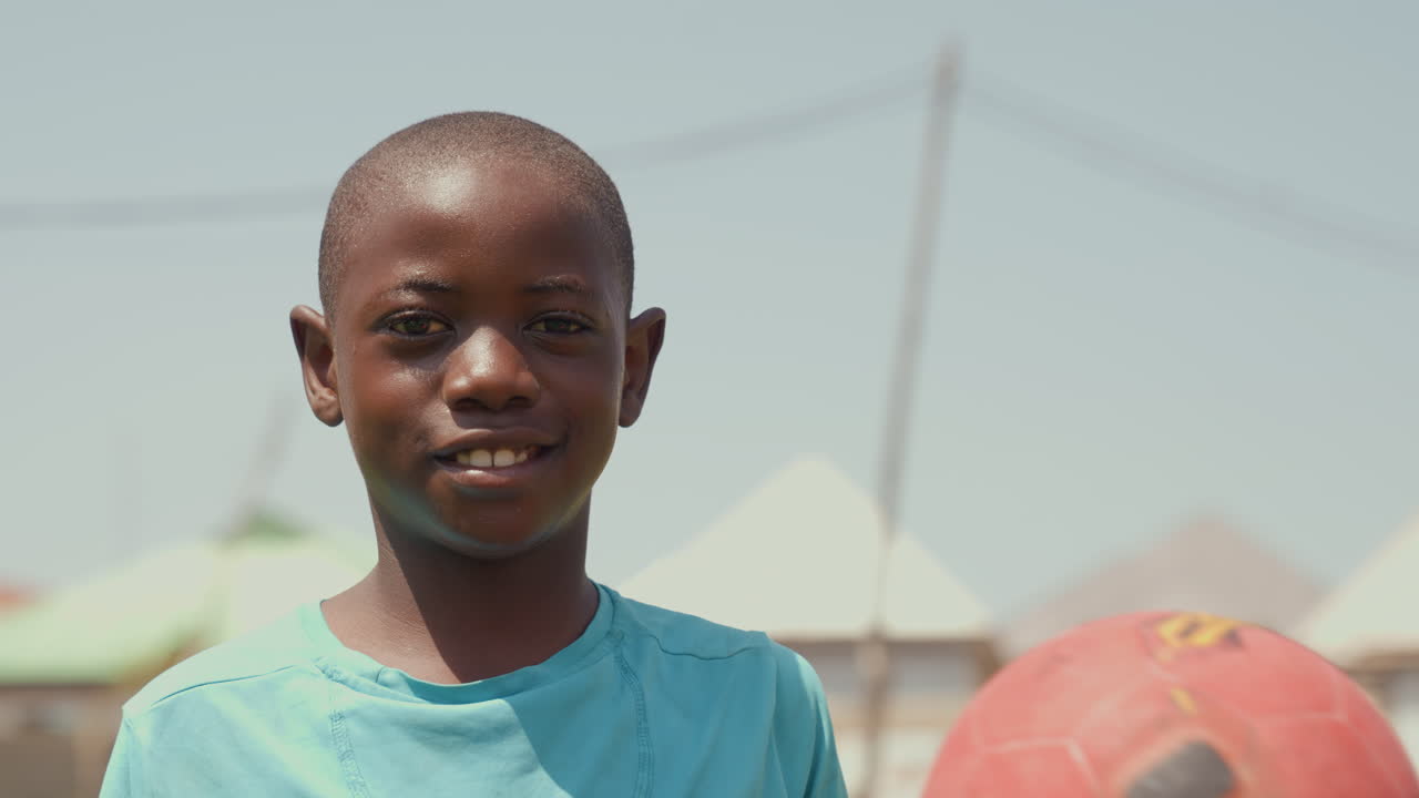 Ragazzo nero che tiene un pallone da calcio rosso, scena di quartiere illuminata dal sole con maglietta turchese e sorriso fiducioso, un primo piano spontaneo cattura l'energia giovanile, gioco spontaneo, luce estiva, campo della comunità.