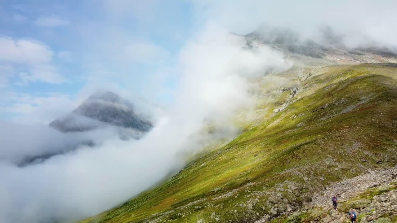 Aerial drone footage flying over green grassy and rocky Sweden hills with low hanging white fluffy clouds while two people are seen hiking and walking forward on the rural muddy and rocky nature road.