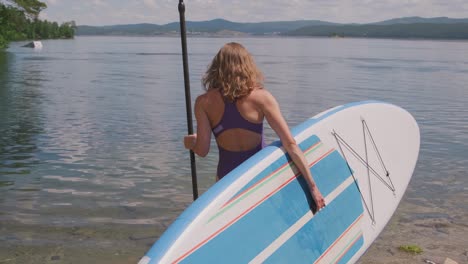 vista posteriore della ragazza bionda in costume da bagno che porta il paddle board a riva e inizia a pagaiare