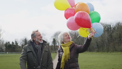 Liebendes-älteres-Paar-Mit-Luftballons,-Das-Gemeinsam-Einen-Herbst--Oder-Winterspaziergang-Durch-Den-Park-Genießt