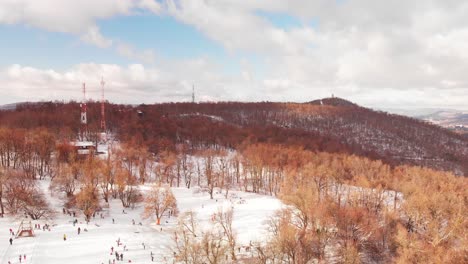 Aerial shot of a drone hovering above a beautiful winter land on the snowy hills with people sledging, hiking and playing in the background