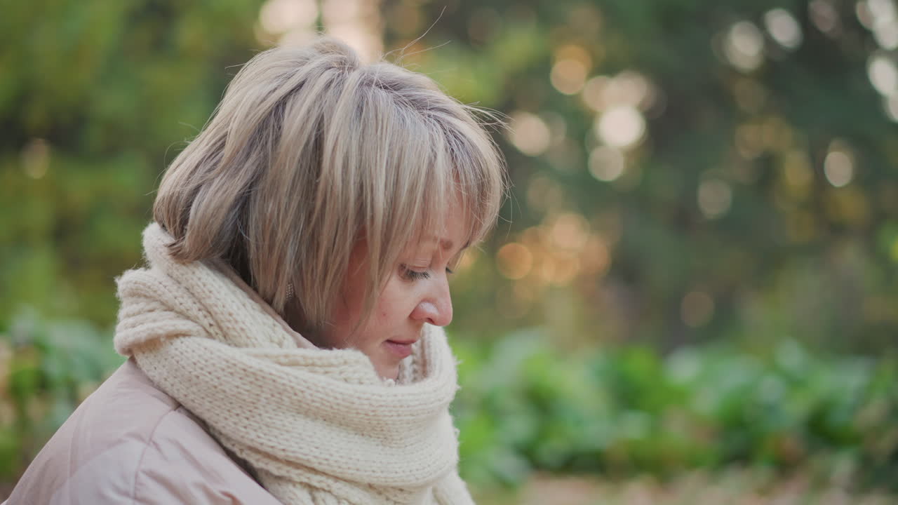 close up side view of mature woman in warm scarf with soft short hair focusing calmly on something out of frame, gentle bokeh effect in autumn forest setting with muted background