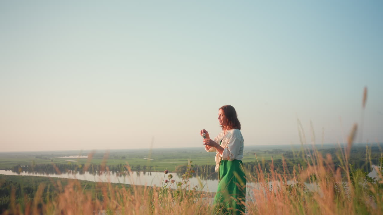 Fair skinned woman in flowing green dress standing on cliff edge near river valley blowing iridescent soap bubble into warm evening sky, tall grasses swaying around her as soft sunset light bathes