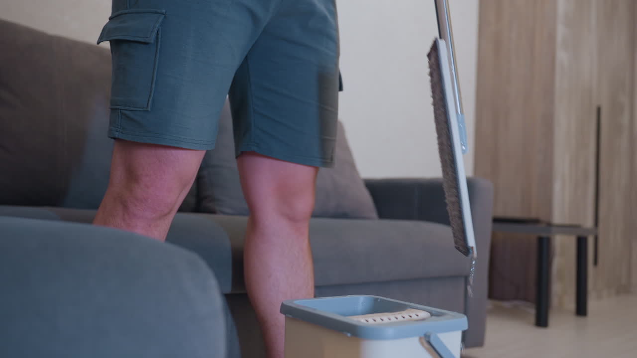 Low angle view of boy in orange shirt with headphones around neck standing indoors holding mop handle with visible water stain on shirt showing casual household cleaning
