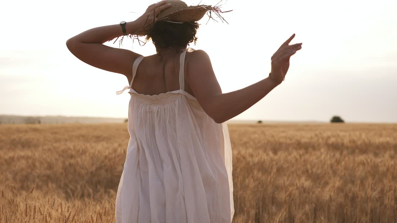 mujer joven corriendo en el campo de trigo mientras sostiene su sombrero de paja. el sol se esconde en el cielo despejado. día de verano. vista rara