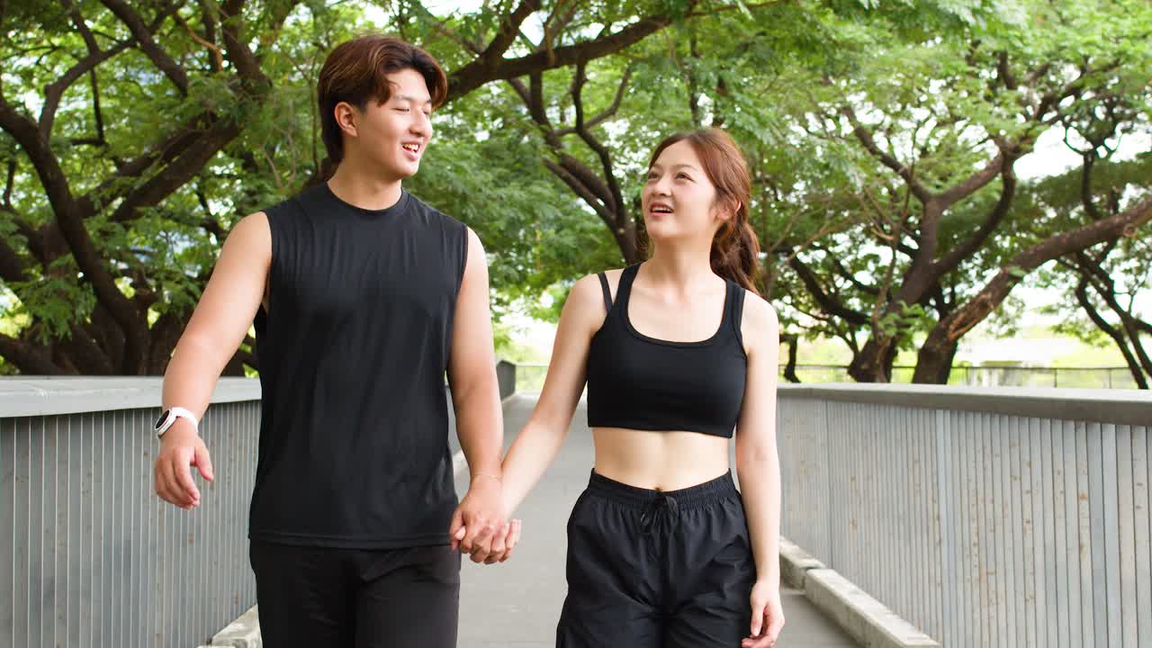 검은색 운동복을 입은 웃는 아시아인 커플이 가로수가 늘어선 도시 길을 따라 손을 잡고 파워 워킹한다.