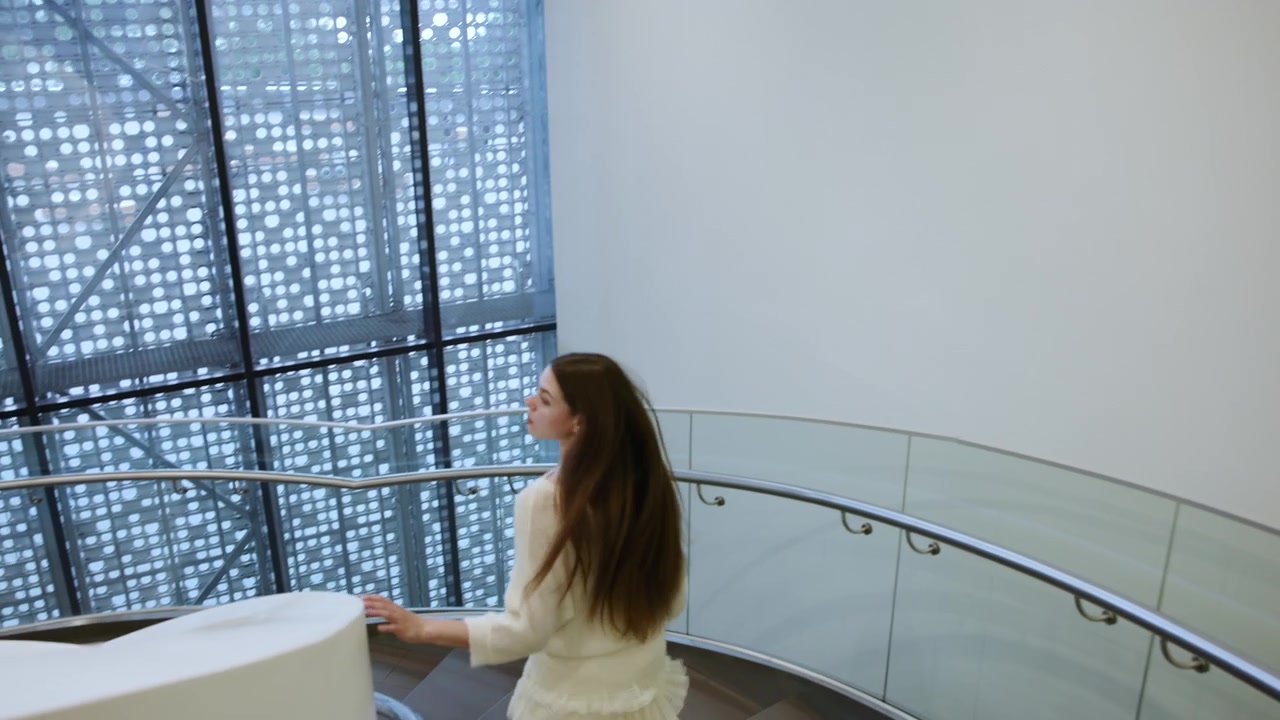 Mulher descendo uma escada em espiral curva em um interior moderno, segurando o corrimão de aço inoxidável para se equilibrar, com cabelos castanhos longos, blusa de tricô e saia com babados, vista de três quartos por trás