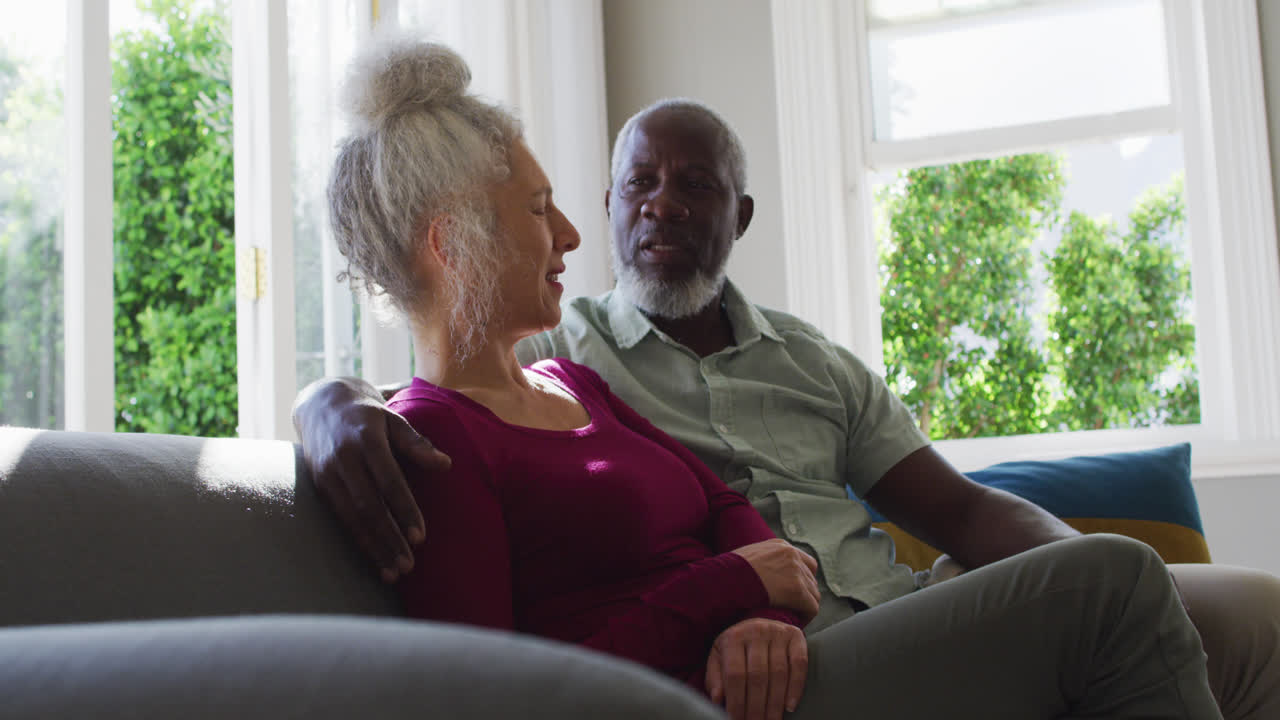 Mixed race senior couple taking to each other while sitting on the couch at home