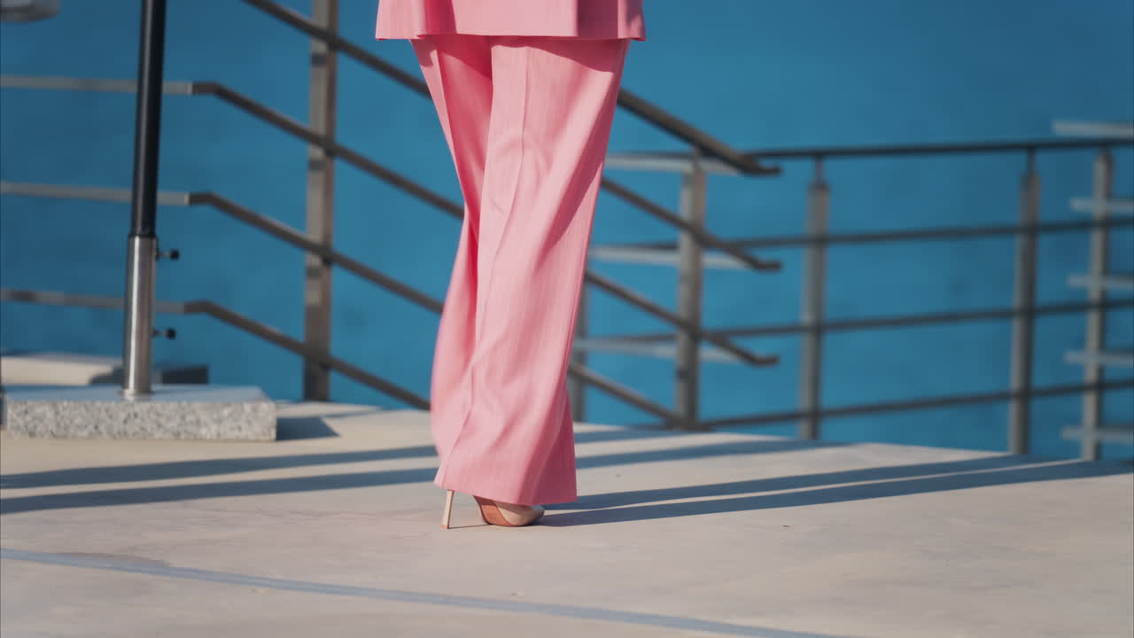 Woman in a pink suit with tan high heels walking on a scenic terrace with a view of a calm body of water