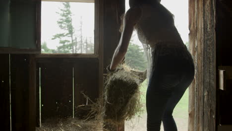 stylish young cowgirl in a barn tosses straw down to her horse