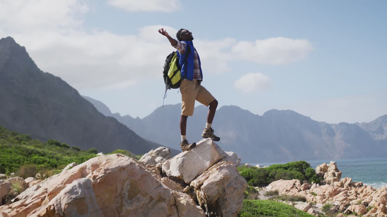 hombre afroamericano de pie en la roca con los brazos abiertos mientras trekking en las montañas