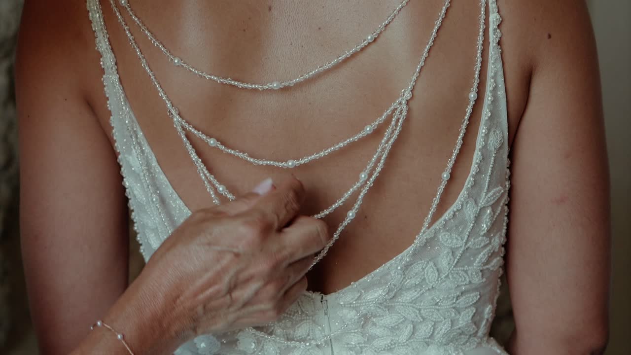 Primer plano de una novia abrochándose un vestido de novia de encaje adornado con detalles de cadena de perlas en la espalda