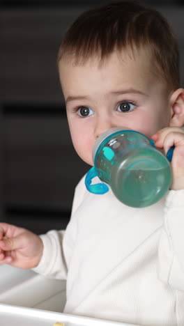 little toddler sitting at the feeding table reaches hand for the bottle. mom's hand gives a bottle, kid drinks water and puts it on the table. close up. vertical video
