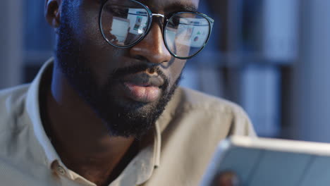 close up view of young smiled man in glasses taping and scrolling on the tablet in the office at night