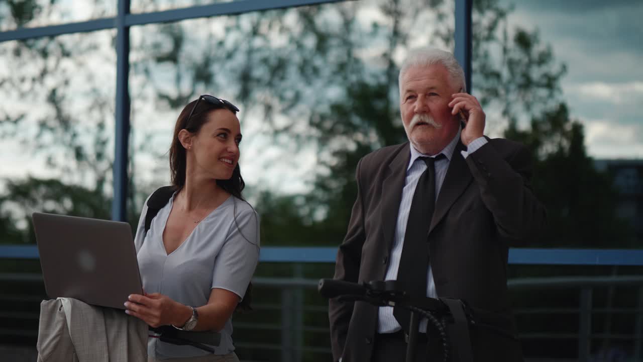 hombre viejo, el director de la compañía, responde el teléfono inteligente mientras está de pie al lado de una trabajadora y un scooter eléctrico