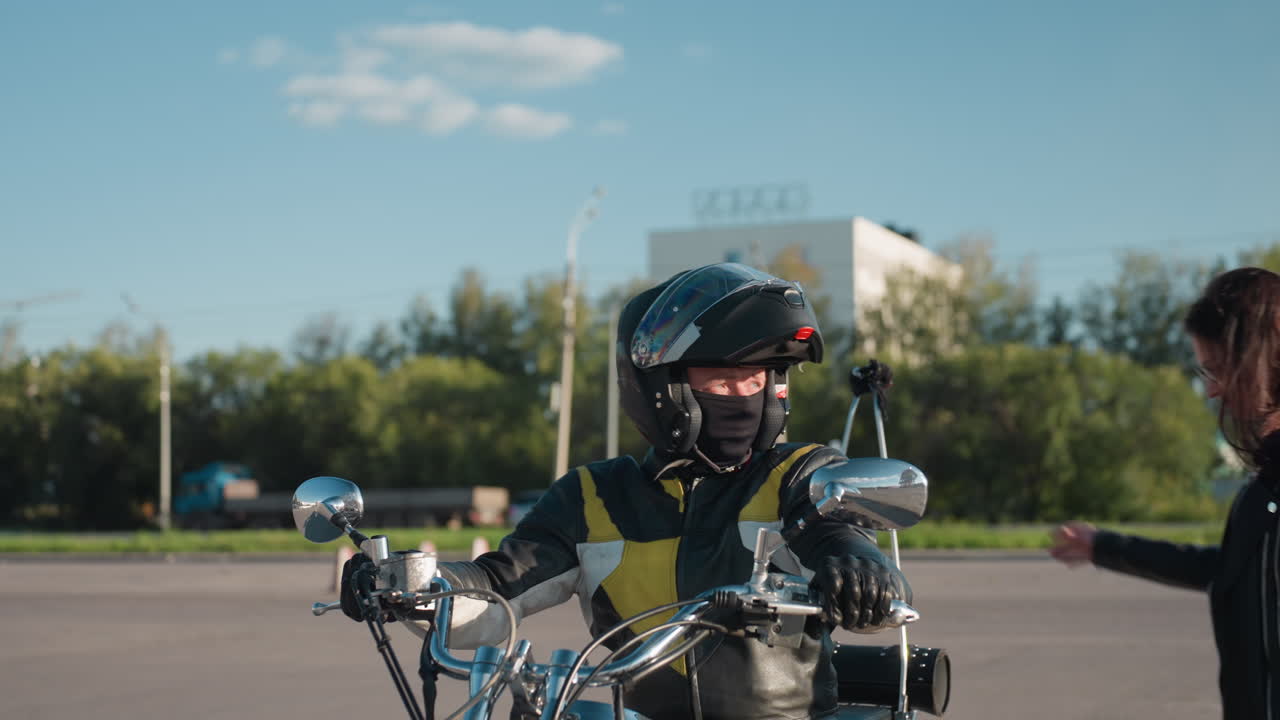 Hand of person raised to stop motorcyclist wearing helmet and mask on city street, rider halts on classic bike under clear sky with urban buildings, vehicles and trees in background