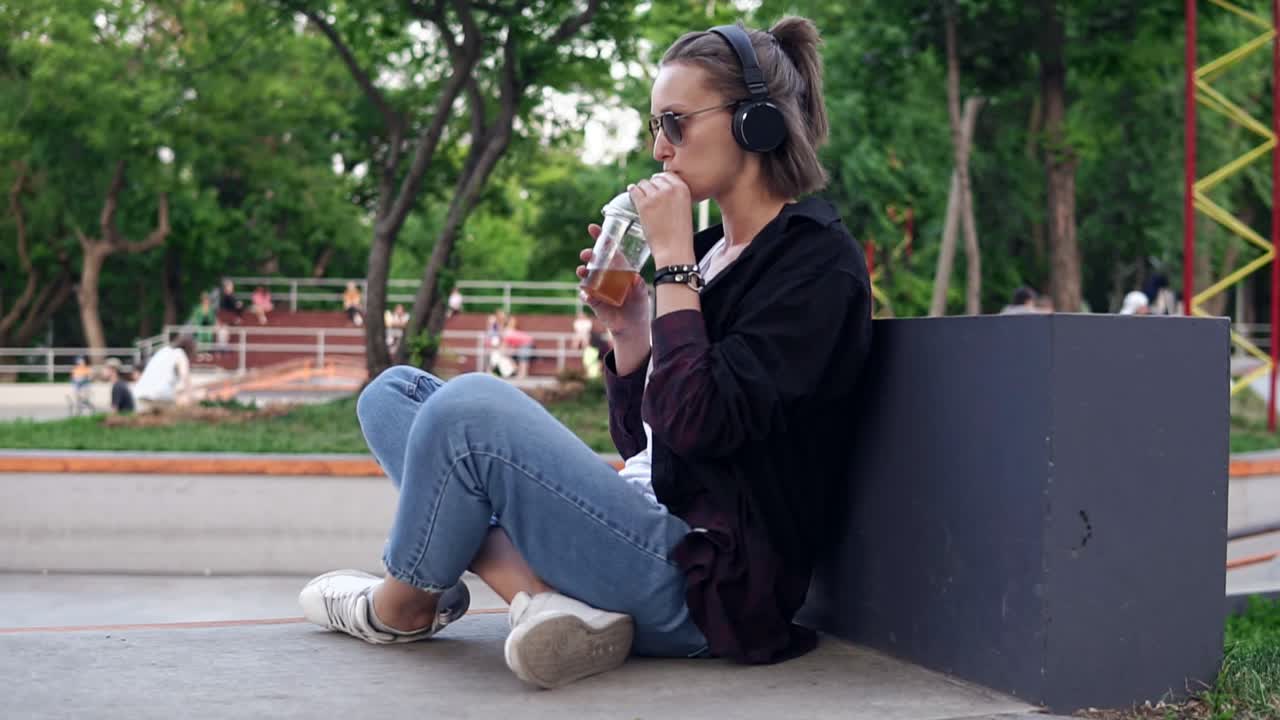 chica joven con estilo se sienta en un parque en el suelo con las piernas cruzadas. bebiendo un refresco. una mujer con gafas de sol y