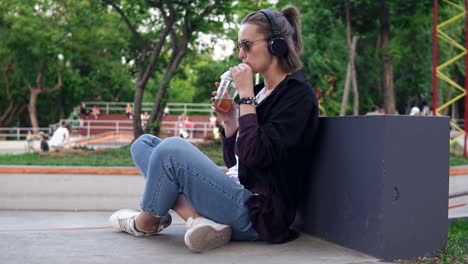 stylish, young girl sits in a park on the floor with her legs crossed. sipping a soft drink. a woman with sunglasses and