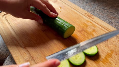 close up of woman's hands slicing knife into fresh cucumber on wood cutting board