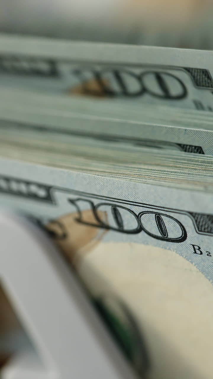 New hundred dollar bills quickly falling on the holder of machine. Close up. Financial equipment at work. Blurred backdrop. Vertical video