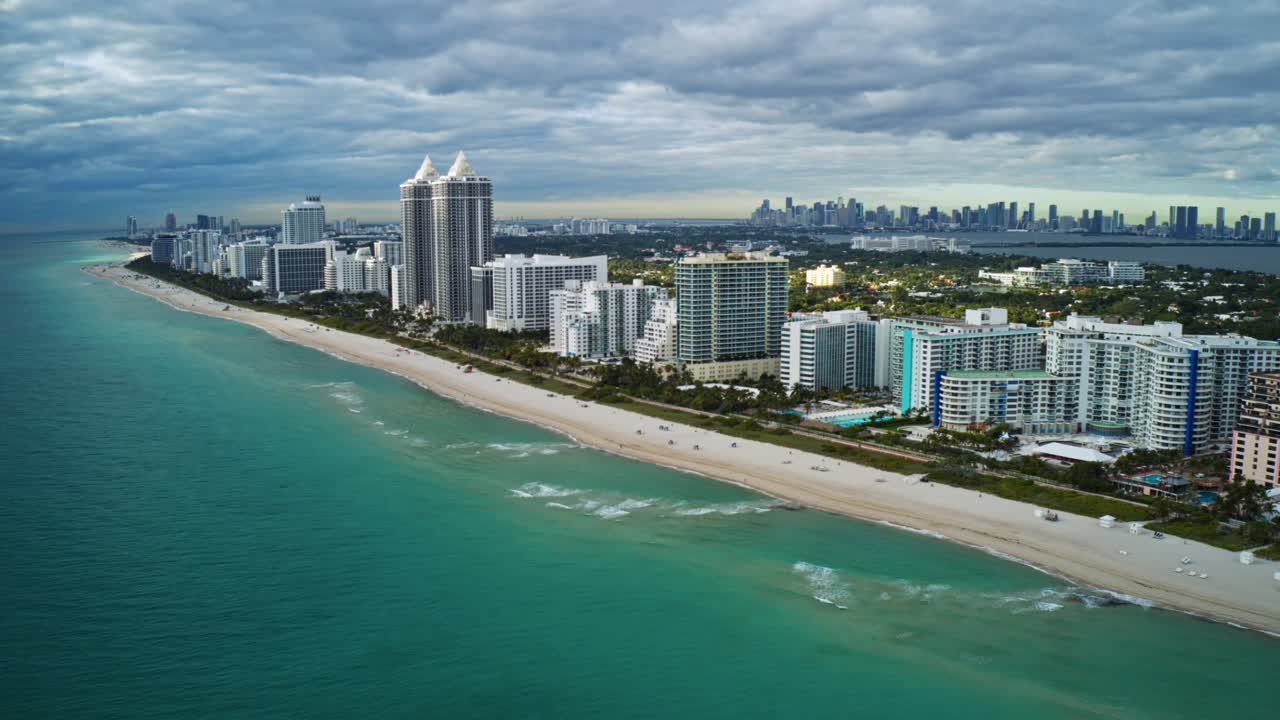 Tall oceanfront condos line the sandy shoreline of North Beach as Miami’s skyline rises in the distance, framed by turquoise Atlantic water and textured cloud cover along Florida’s southeast coast
