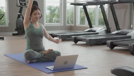 young asian woman doing yoga with sitting and stretch muscles hands at fitness gym, female training practicing workout with exercise while watching laptop computer for bodycare at home, sport concept.