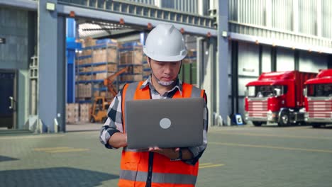 asian male engineer with safety helmet working on a laptop while standing , outside of logistics distributions warehouse