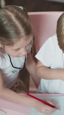 caring sister draws picture for little brother sitting at desk at home. little girl shows exercise to junior boy playing school together in children room