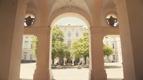 Arched-walkway-leading-to-a-sunny-courtyard-with-trees-and-relaxed-pedestrians,-warm-tone