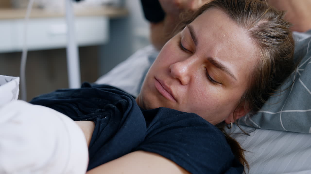 First breastfeeding of a newborn right after birth. Woman lies in bed feeding her tiny child. Close up.