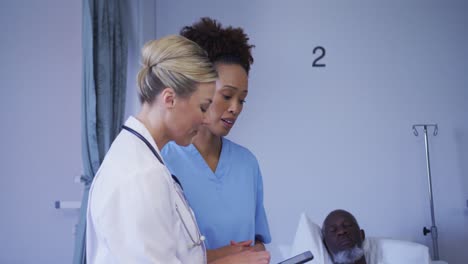 diverse female doctors looking at tablet standing near patient in hospital bed