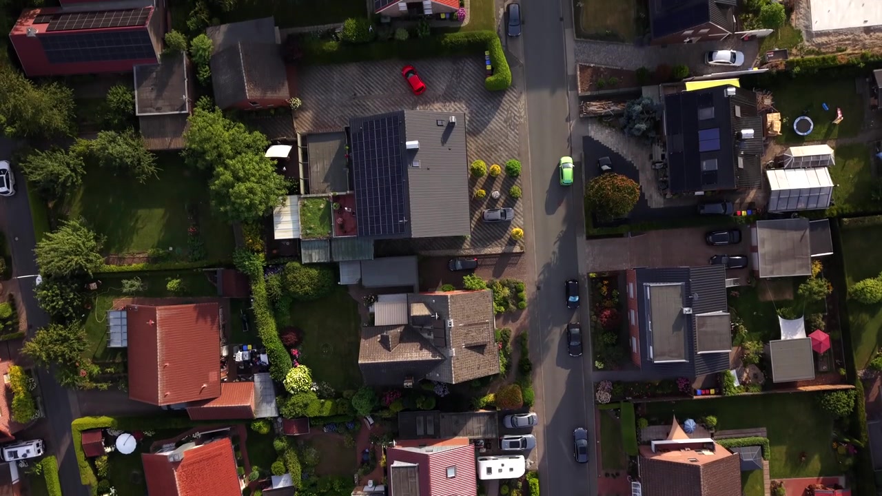 Tetti di tegole rosse si raggruppano attorno a una stretta strada con incroci fiancheggiata da auto parcheggiate, siepi e giardini curati che circondano le case in un tranquillo quartiere tedesco. Sorvolo aereo con drone dall'alto