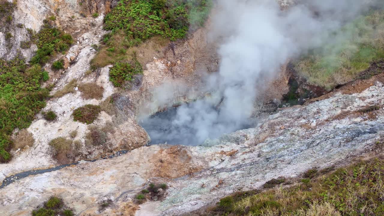 Top view of a sulfur crater releasing white smoke from geothermal activity, located in a mountain area with a mix of rocks and greenery. Sikidang Crater, Dieng, Indonesia