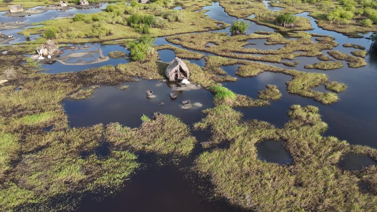 Spectacular bird's-eye view of the coastal marshlands in Ouidah, Benin. The drone shot reveals traditional thatched-roof huts surrounded by lush green vegetation and calm inland waters