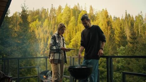 A-blonde-girl-helps-a-brunette-guy-cook-meat-on-the-grill-on-the-balcony-of-a-country-house-against-the-backdrop-of-a-coniferous-forest