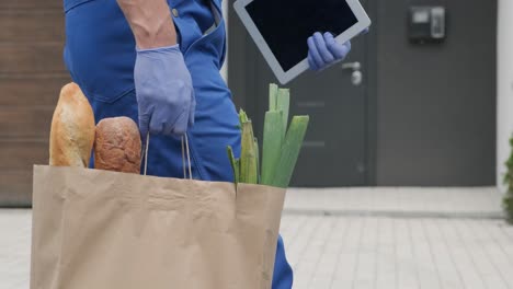 junger kurier mit schutzmaske und handschuhen liefert ein einkaufspaket mit nahrungsmitteln direkt an einen kunden zu hause mit sicherheit. lebensmittellieferung, hauslieferung in quarantäne