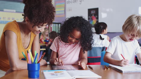 female elementary school teacher sitting at desk helping pupils in classroom