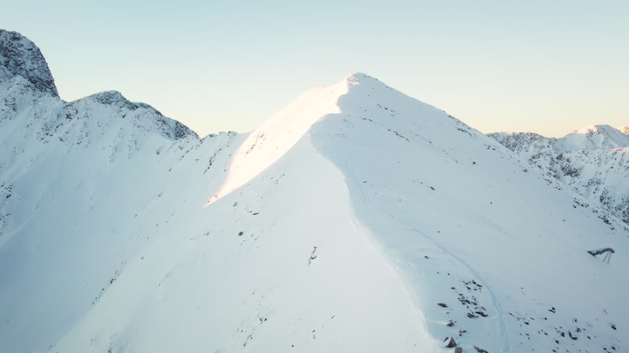 Drone ascends sunlit slope to mountain summit, revealing majestic snowy peaks behind. Filmed in Andorra.