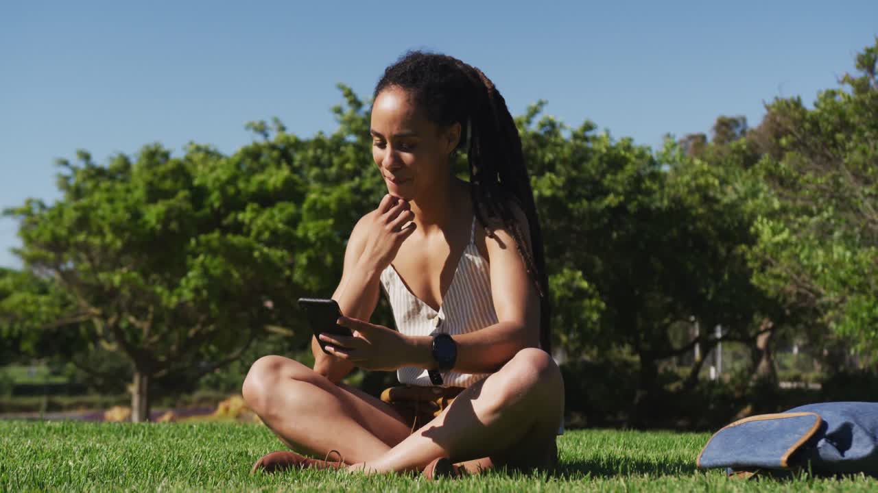 African american woman sitting on grass using smartphone in park