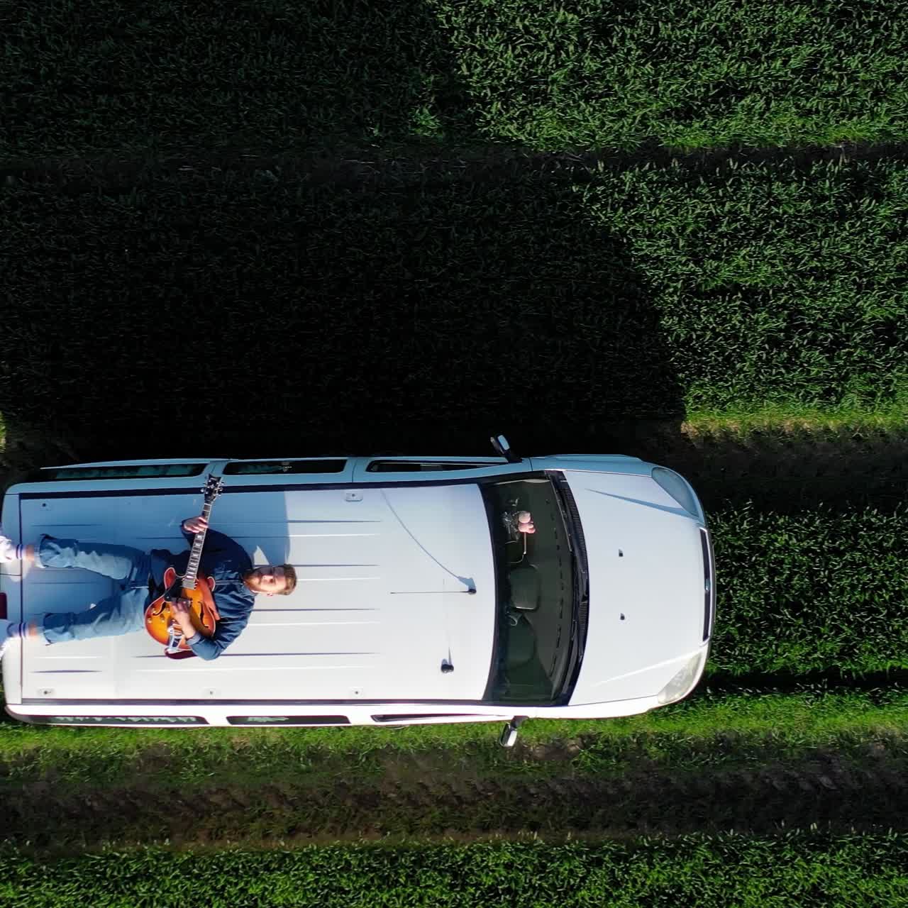 Extreme driving on a roof of a car. Bearded guy lying on a moving car and playing the guitar on green field background. Aerial view