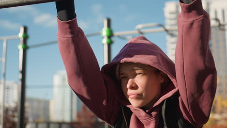 fitness instructor gripping pullup bar under bright sunlight, hoodie clad athlete showing resilience, gaze squinting against glare while performing outdoor exercise on urban workout station