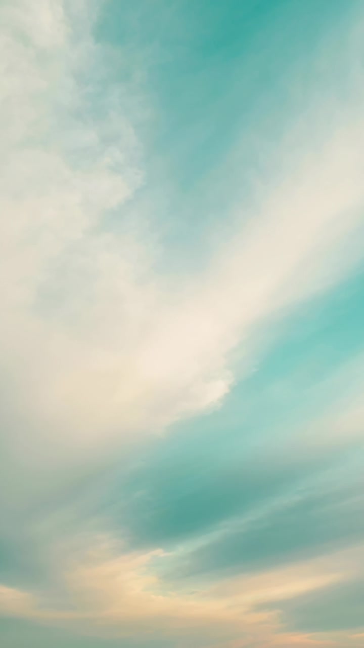 Vertical video: Drifting cloud layers responding to sunrise light over open sky, warming teal tones