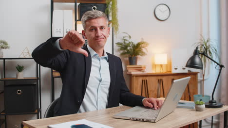 upset young caucasian freelancer in suit using laptop and showing thumb down at home office table