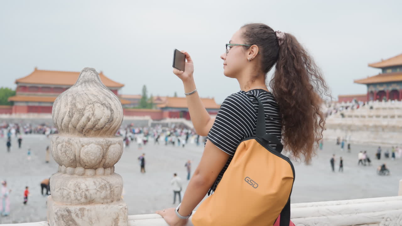 Donna di colore backpacker nella piazza della Città Proibita, con zaino giallo e occhiali, che alza lo smartphone per una foto grandangolare. Folla vivace e tetti di palazzo, energia da blogger che cattura persone e panorama.
