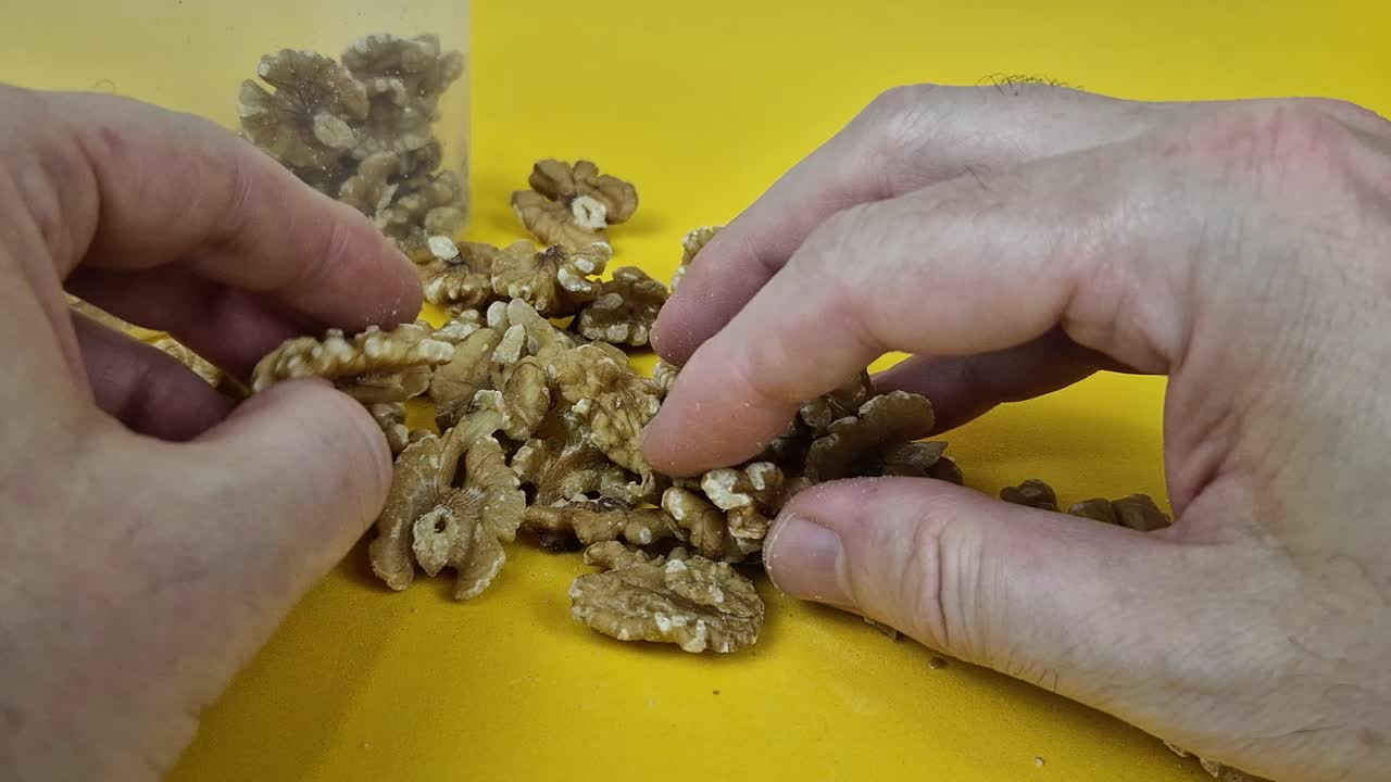 Detailed 4K macro shot of shelled walnut halves and pieces scattered on a vibrant yellow surface.A person's hands can be seen interacting with the nuts