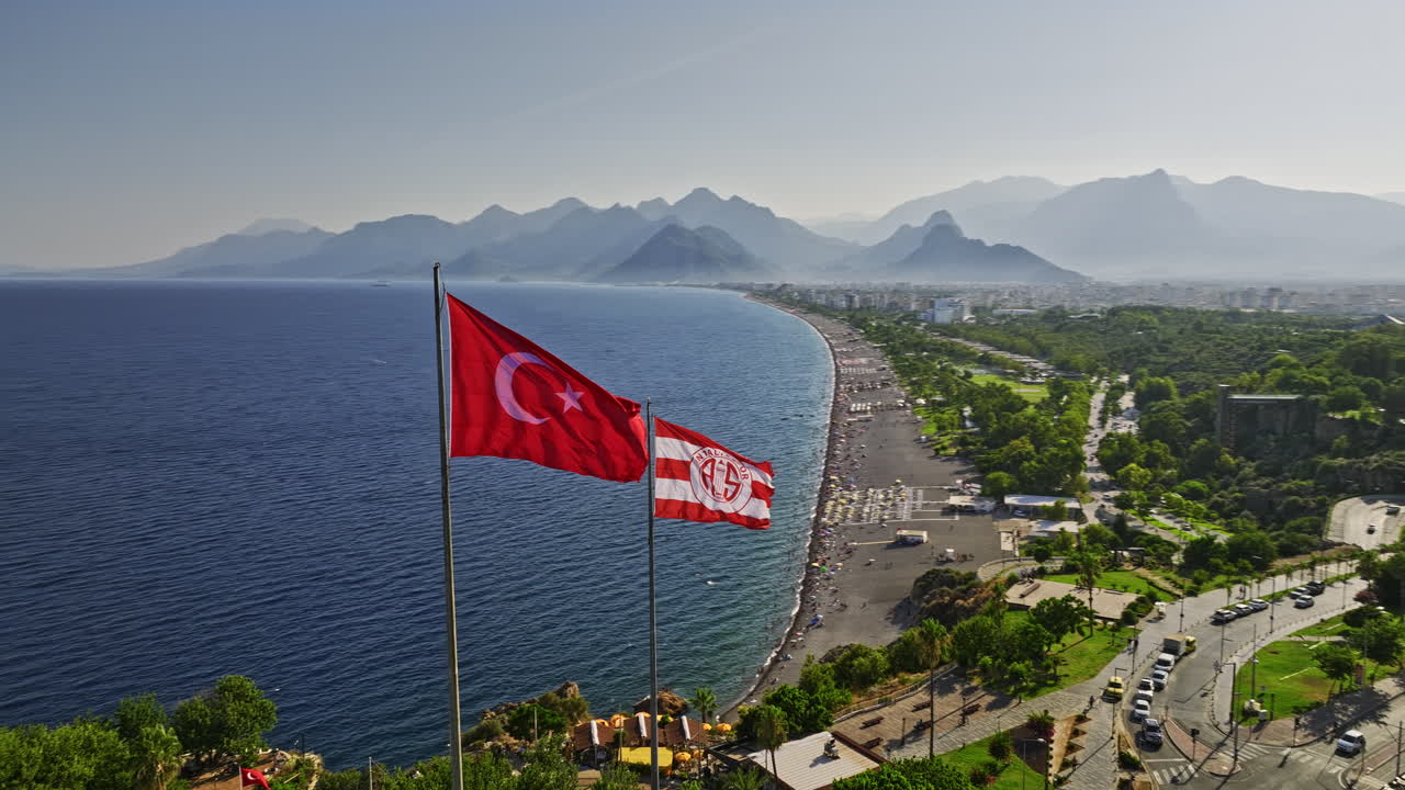 Antalya Turecko Letecký v13 panoramatický pohled z dronu létajícího kolem turecké národní vlajky a vlajek Antalyasporu ve větru meltem, zachycující pláž Konyaaltı s výhledem na siluetu hor – natočeno dronem Mavic 3 Cine – červenec 2022.